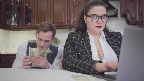 年轻自信的胖女人 戴着眼镜坐在厨房的桌子上 拿着笔记本电脑 男人坐在女士后面数钱 — 图库视频影像