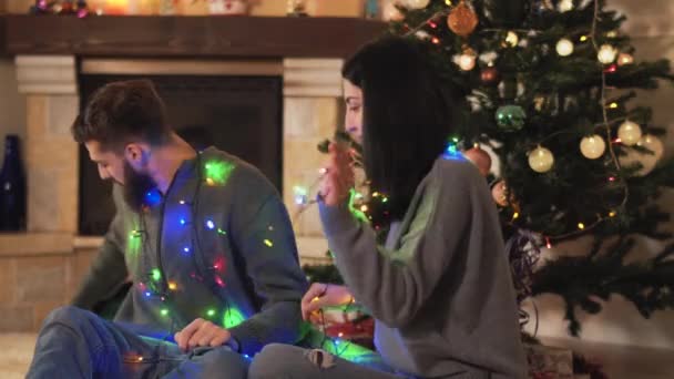 男人和女人坐在圣诞树旁 脖子上挂着花环 男人送给女人礼物 — 图库视频影像
