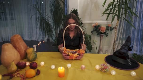万圣节 漂亮的巫婆手里拿着一篮子糖果 南瓜和水果旁边桌子上的女巫帽 万圣节假期录像 — 图库视频影像