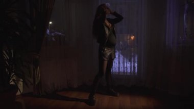 Kısa deri şortlu güzel hamile kadın karanlık odanın penceresinin yanında duruyor. Modern elbiseli bayan dışarı çıkacak, video. 