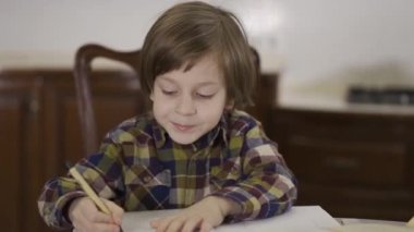Kareli tişörtlü şirin çocuk evde, masada oturmuş ödevini yapıyor. Okul çocuğu defterine resim çiziyor. Kamera sağa sola hareket ediyor