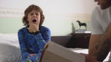Sevimli bir çocuk büyük bir yatakta oturup gözlerini ovuşturuyor çünkü erkek kardeşi onun için kitaptan çok yorgun düşmüş..