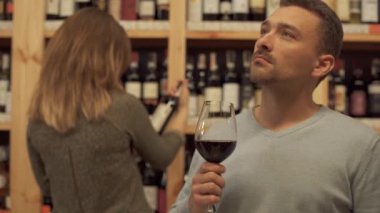 İçki dükkanında elinde bir bardak kırmızı şarapla duran yakışıklı adamın portresi. Arkadaki kadın şarap şişesini seçiyor. Bol alkollü bir yer..