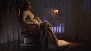 Uzun saçlı güzel hamile kadın karanlık odada sallanan sandalyede oturuyor. Bayan karnını okşa. Kadının bacakları battaniyeyle örtülmüş. Kadın yumuşak oyuncağa sarılıyor, video... 