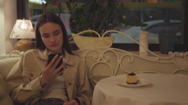 Restorandaki masada oturan cep telefonuyla mesajlaşan güzel kız. Lezzetli kek dilimi masanın üzerinde. Kafenin içi rahattır.