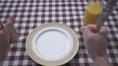 Adamın eli masaya yumruklarıyla vuruyor, yemek istiyor, çatal ve bıçağı yakından tutuyor. Boş tabak ve portakal suyu bardağı kareli masa örtüsüyle masada. Sahanda yumurta tabağa düşer. Erkeğin kahvaltısı. Beslenme kavramı. 