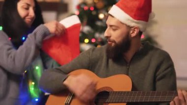 Gitar çalan ve Noel ağacının yanında şarkı söyleyen bir adam, şapka takan bir kadın, video. 