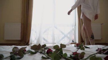 Genç bir kadının kısa şeffaf gömleğiyle odada yürüdüğü ve arka plandaki pencereye geldiği bir video. Kız tülleri geri çeker ve dışarı bakar. Kırmızı güller ön planda, yatağın üstünde.