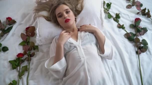 透明パジャマ姿でベッドに横になって明るいメイクの若い女性の動画 赤いバラは 遊び心のある気分で彼女の体に触れて 女性の体の近くのベッドにあります — ストック動画