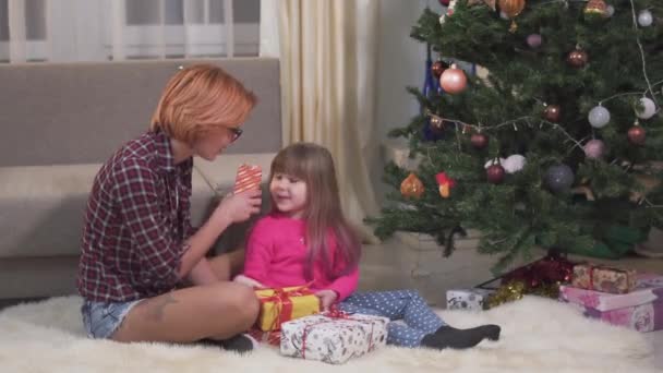 面白い女の子と彼女の母親はクリスマスツリーの下に座って 手にいくつかの現在のボックスを保持し 短い赤い髪と彼女の子供を持つ若い女性は休日前に一緒に楽しみを持っています — ストック動画