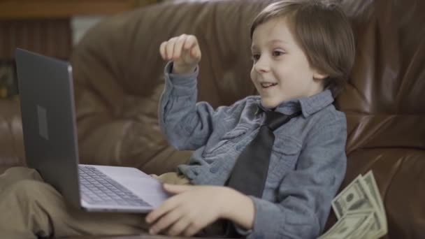 穿着牛仔裤夹克的小男孩坐在皮革沙发上 在笔记本电脑上玩耍 美元掉在沙发上 孩子在笑 现代儿童的闲暇时间 有钱父母的儿子真可爱 慢动作 — 图库视频影像