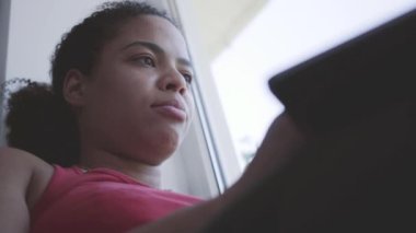 Pencere eşiğinde oturan ve dizüstü bilgisayarda yazan Afrikalı Amerikalı kadının videosu. Mutlu kız evde yalnız başına dinleniyor.