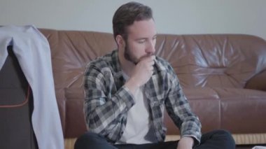 Yorgun sakallı adamın yerde oturup bavullarını toplarken videosu. Yolculuk için katlanacak bir sürü giysi ve ayakkabı var.