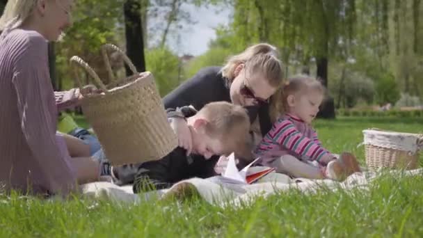 春天的一天 两个带着孩子的年轻母亲坐在大自然的绿意盎然的公园里的毛毯上 妈妈和儿子 妈妈和女儿在野餐 白色绒毛狗躺在附近 家庭间友谊的概念 — 图库视频影像
