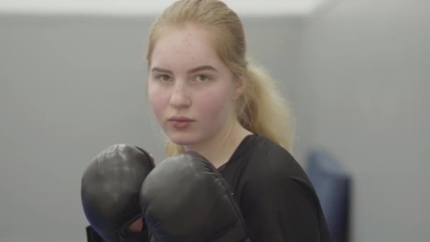一个不自信的金发年轻女子戴着拳击手套 模仿躲躲闪闪的拳打脚踢在体育馆里的肖像 可爱的新手跆拳道手 武术的概念 — 图库视频影像