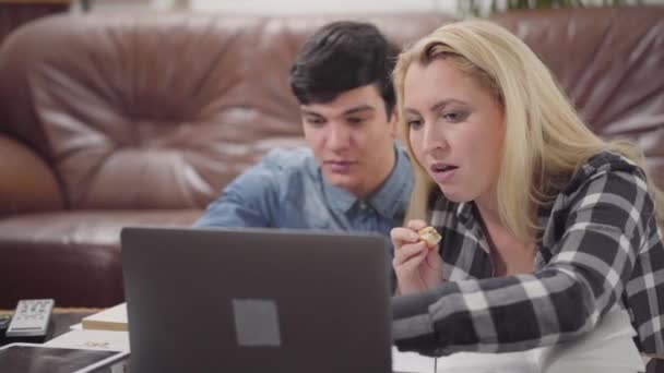 若いカップルは自宅でノートパソコンの前に座っている 一緒にラップトップで働いているブルネットの男とブロンドの女性の肖像画 女の子はケーキを食べています 背景に革のソファ — ストック動画