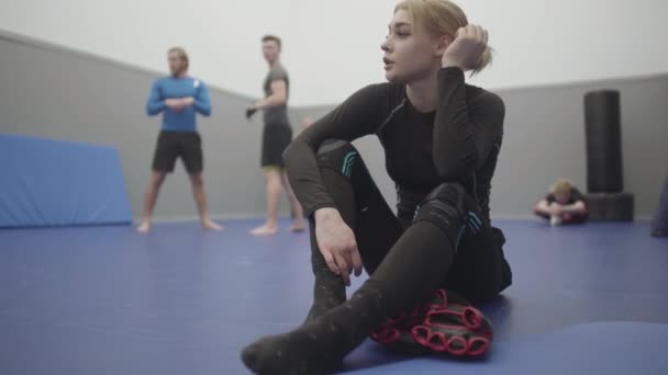 那位漂亮的女运动员坐在体育馆的地板上 训练完瓶装水后 休息了一下 金发女士在背景音乐中拳打脚踢 武术的概念 — 图库视频影像