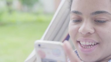 Genç Afrikalı Amerikalı kadın hamakta oturuyor, bahçede dinleniyor ve akıllı telefon ve video kullanıyor. 