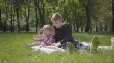 Parkta battaniyeye oturmuş, yanında oturan pembe elbiseli kız kardeşine kitap okuyan sarışın bir çocuk. Kız hikayeyle ilgilenmiyor. Oynamak istiyor. Çocuklar piknikte
