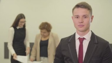 Kadın meslektaşları arka planda evraklarla çalışırken yakışıklı, kendine güvenen, gülümseyen genç bir adamın videosu. 