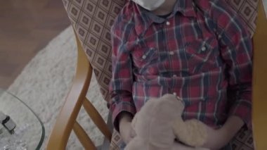 Tanınmayan çocuk koltukta sallanıyor, elinde yumuşak bir oyuncak, haplarla dolu bir masa, video