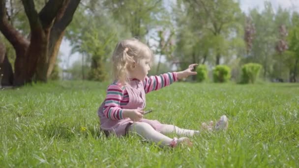 かわいい女の子は 公園内の草の上に座って 一人で遊んで 小さな指を指しています 屋外で休んでいる気をつけてください 健康的なライフスタイル 自然とのつながり — ストック動画