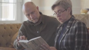 Yaşlı bir çift, kadın ve erkek, kanepede oturmuş kitap okuyorlar ve birbirlerine gülümsüyorlar. Erkek keldir ve dişinin kısa gri saçları vardır. Mutlu sevgi dolu aile.