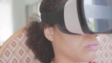 Sanal gerçeklik kulaklığı takan Afro-Amerikalı bir kadının evdeki koltukta oturduğu video. Video oyunu oynayan kız. 