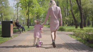 Genç anne, parktaki kızla birlikte kızının scooterına biniyor. Arkadan bak. Çocuk başını çevirir ve kameraya bakar. Aktif yaşam tarzı, boş zaman, tasasız bir çocukluk.