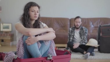 Gücenmiş bir kadın bavulda oturuyor, mutlu bir koca bavulunda eşyalarını topluyor, video çekiyor. 