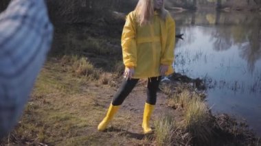 Sarı paltolu ve lastik çizmeli, ormanın yakınında poz veren sarışın genç bir kadın. Adam cep telefonuyla fotoğrafını çekiyor..