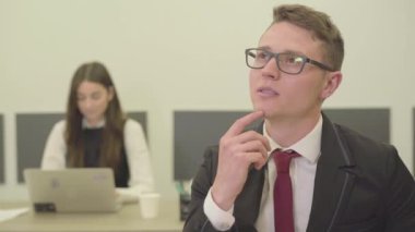 Ofiste ön planda oturan düşünceli genç bir adamın videosu. Resmi olarak bayan meslektaşı arka planda dizüstü bilgisayarla çalışırken.