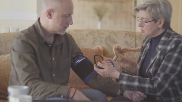 年长的夫妇坐在沙发上 灰白短发的女人用体能计测量男人的血压 — 图库视频影像