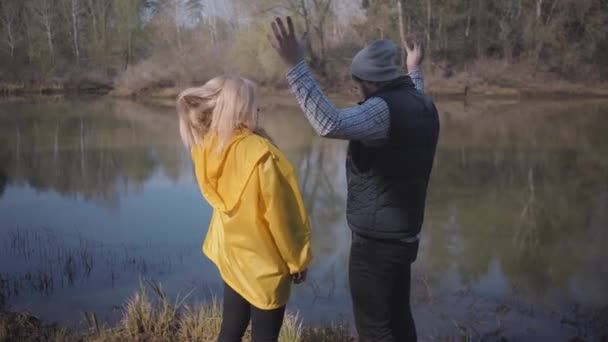 一对时髦的夫妇在森林里看着河流 穿着背心 太阳镜的男人 穿着黄色外套和靴子的金发女人 — 图库视频影像