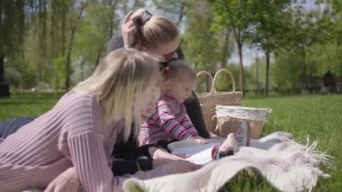 Parktaki battaniyenin üzerinde çocuklarıyla birlikte iki genç anne. Genç sarışın çocuk yalan söylüyor ve kitap okuyor. Pembe elbiseli şirin küçük kız yakında oynuyor. Çocuklarla piknik.