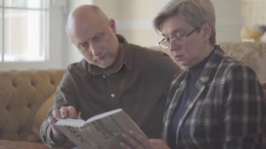 Yaşlı bir çift, kadın ve erkek, kanepede oturmuş kitap okuyorlar ve birbirlerine gülümsüyorlar. Erkek keldir ve dişinin kısa gri saçları vardır. Mutlu sevgi dolu aile