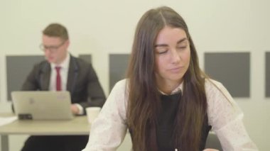 Resmi olarak giyinmiş düşünceli bir kadının ofiste ön planda otururken videosu. Ofiste dizüstü bilgisayar kullanan bir adam. 