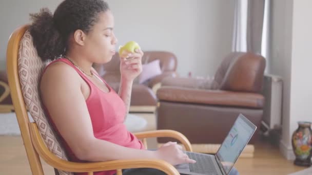 非洲裔美国妇女坐在扶手椅上分析笔记本电脑上的图表 同时吃青苹果的视频 — 图库视频影像