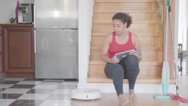 漂亮的非洲裔美国女人坐在大房子一楼的楼梯上 用平板电脑发短信 现代机器人真空吸尘器 — 图库视频影像