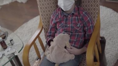 Çocuk koltukta sallanıyor, elinde yumuşak bir oyuncak, haplarla dolu bir masa, video