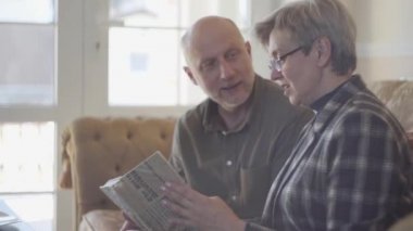 Yaşlı bir çift, kadın ve erkek, kanepede oturmuş kitap okuyorlar ve birbirlerine gülümsüyorlar. Erkek keldir ve dişinin kısa gri saçları vardır. Mutlu sevgi dolu aile.