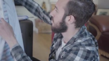 Yorgun sakallı adam yerde oturuyor ve bavullarını topluyor. Evde bir sürü kumaş ve ayakkabı var. 
