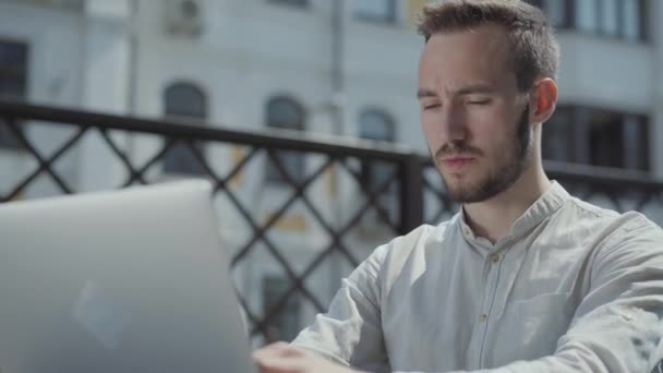 髭を生やした若い男がノートパソコンの前のテラスのテーブルに座って働いていた フリーランスの概念 遠くの仕事 屋外でビジネスをしてる男ビデオ — ストック動画