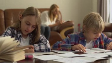 Anneleri arka planda otururken, evde ders çalışan sevimli küçük kız ve erkek çocukları. Çocuklar ödevlerini evde birlikte yapıyorlar.