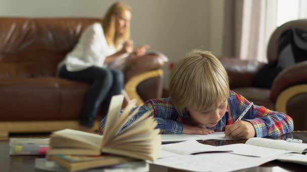 母親がソファの上に座っている間 かわいい男の子は前景の自宅のノートブックに情報を書き留めています — ストック動画
