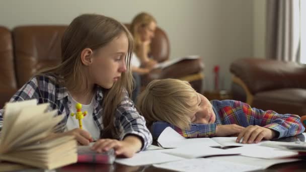 母親が背景に座っている間 かわいい男の子と女の子は前景に家で勉強します 兄は眠りに落ち 妹は彼を目覚めさせた 子供たちは一緒に宿題をする — ストック動画