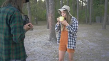 Genç adam, ormanın arka planında bir fincan çay ya da kahveyle güzel bir hipster kızı poz verirken küçük bir kamera ile video çekiyor.