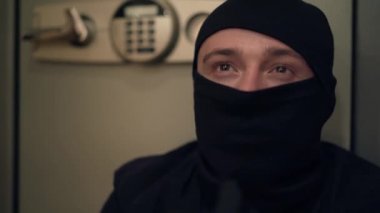 Close-up of young Caucasian thief in black clothes and balaclava looking at camera. The guy stealing things from safe. Concept of crime, criminal, offense against the law