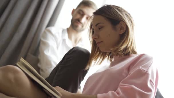 年轻漂亮的夫妇正坐在家里的窗边 一个女孩正在给她的男朋友或丈夫读书 — 图库视频影像
