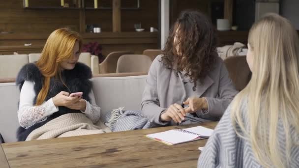 三个白人女朋友坐在咖啡厅的桌旁 一个女孩拿着电话 另两个讨论写在笔记本上的项目 年轻女人一起在室内放松 秋天的闲暇 友好会议 — 图库视频影像